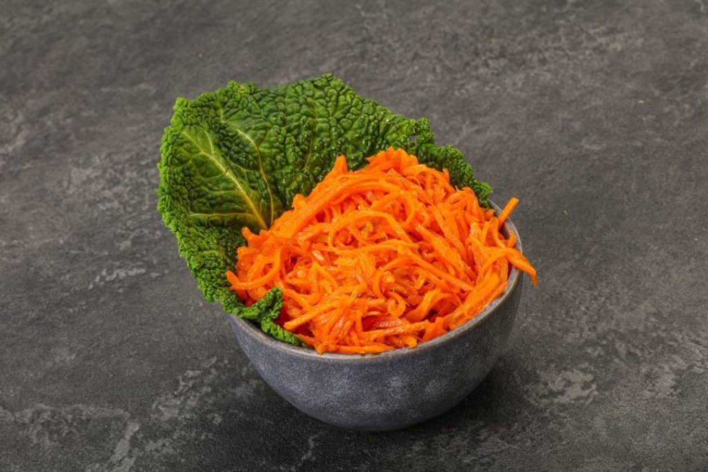 Морква по-корейськи — це салат, який давно став частиною домашньої кухні, хоча має зовсім не українське походження. Його готують на свята, додають до м’ясних страв і подають як самостійну закуску. Якщо ви шукаєте, як приготувати моркву по-корейськи так, щоб вона була хрумкою, ароматною та збалансованою за смаком, цей матеріал стане покроковим орієнтиром. Ми розберемо покроковий рецепт, пояснимо роль спецій і покажемо, як змінювати гостроту без втрати характеру страви. Також ви дізнаєтесь, як зберігати салат і чи підходить морква по-корейськи на зиму. У тексті є практичні поради, приклади та варіанти подачі. Структура побудована так, щоб ви могли не просто повторити рецепт моркви, а зрозуміти логіку маринаду. Це дозволить готувати стабільно смачно навіть без вагів і точних мір. Що таке морква по-корейськи Морква по-корейськи — це маринована морква з додаванням оцту, спецій та олії, яка має яскравий аромат і легку гостроту. Хоча назва натякає на корейське походження, страва виникла як адаптація корейських салатів до доступних інгредієнтів. Морква корейська готується просто, але вимагає розуміння балансу кислоти, солі та спецій. У домашніх умовах важливо правильно нарізати овоч і дати йому час промаринуватися. Корейська морква в домашніх умовах часто відрізняється від магазинної більш натуральним смаком і свіжістю. Салат підходить як гарнір до м’яса, картоплі або рису. Його готують заздалегідь, адже смак розкривається не одразу. Саме тому запит “морква по-корейськи” стабільно популярний серед тих, хто шукає простий і яскравий салат. Історія та походження Історія страви пов’язана з корейською діаспорою, яка адаптувала традиційні рецепти до доступних продуктів. Замість рідкісних овочів використовували моркву, що була доступною та дешевою. Так з’явилася морква по-корейськи у сучасному вигляді. Рецепт моркви швидко поширився завдяки простоті приготування. Страва не потребує термічної обробки, лише правильного маринування. З часом з’явилися варіації з різними спеціями для моркви. У кожному регіоні додають щось своє — більше часнику, гострого перцю або цукру. Сьогодні морква по-корейськи сприймається як класична закуска до багатьох страв. Чому вона така популярна Популярність пояснюється простотою та яскравим смаком. Якщо ви думаєте, як приготувати моркву смачно без складних інгредієнтів, цей варіант ідеальний. Морква корейська готується з доступних продуктів і не вимагає особливих навичок. Салат довго зберігається в холодильнику, що зручно для зайнятих людей. Його можна зробити гострим або більш м’яким за смаком. Крім того, смакова маринована морква по-корейськи добре поєднується з м’ясними стравами. Вона додає контрасту й текстури до звичних гарнірів. Саме універсальність робить її постійним гостем на столі. Інгредієнти для моркви по-корейськи Щоб морква по-корейськи вийшла збалансованою, важливо правильно підібрати інгредієнти моркви та пропорції. Основу складає свіжа соковита морква, оцет, рослинна олія та спеції для моркви. Деякі рецепти передбачають моркву по-корейськи з оцтом столовим або яблучним, що впливає на смак. Важливо не переборщити з кислотою, щоб салат не став занадто різким. Маринад для моркви по-корейськи повинен бути ароматним, але не домінувати над смаком овоча. Якщо морква солодка, кількість цукру можна зменшити. Рецепт моркви гнучкий, але баланс інгредієнтів має значення. Далі — базовий набір продуктів. Для класичного варіанту підготуйте такі інгредієнти: Свіжа морква — 1 кг Часник — 3–4 зубчики Оцет — 1–2 ст. ложки Рослинна олія — 3–4 ст. ложки Цукор — 1 ст. ложка Сіль — за смаком Червоний перець — за смаком Коріандр мелений Основні компоненти та пропорції Спеції для моркви формують характер страви, тому їх варто додавати поступово. Маринад для моркви по-корейськи повинен містити кислу складову, солодкість і ароматну частину. Рецепт моркви допускає варіації, але базові пропорції краще зберігати. Якщо ви додаєте більше оцту, компенсуйте це невеликою кількістю цукру. Морква по-корейськи з оцтом має виразніший смак, але не повинна бути різкою. Олію часто прогрівають і заливають на спеції, щоб аромат розкрився. Саме цей прийом дозволяє досягти глибшого смаку. Дотримання пропорцій — запорука стабільного результату. Покроковий рецепт моркви по-корейськи Покроковий рецепт допоможе уникнути помилок і отримати хрумку текстуру. Перед тим як приготувати салат, моркву потрібно натерти на спеціальній тертці. Це забезпечить правильну форму й маринування. Далі додаються інгредієнти моркви та спеції. Як приготувати моркву по-корейськи без зайвої гіркоти? Важливо не перетримати оцет і правильно змішати маринад. Смакова маринована морква по-корейськи розкривається через кілька годин. Краще дати їй настоятися мінімум 2–4 години. Так текстура стане м’якою, але залишиться хрумкою. Ось зрозуміла послідовність дій, щоб морква вийшла хрумкою: Натерти моркву на спеціальній тертці. Додати сіль і злегка перемішати. Додати цукор, оцет і спеції. Розігріти олію та залити на спеції. Додати подрібнений часник. Ретельно перемішати. Залишити маринуватися 2–6 годин. Морква по-корейськи найсмачніша після маринування: спеції “розкриваються”, а текстура стає хрумкою й соковитою. Секрети правильної моркви по-корейськи Морква по-корейськи вимагає точності в деталях. Спеції для моркви повинні бути свіжими, інакше аромат буде слабким. Маринад для моркви по-корейськи краще додавати поступово. Якщо ви сумніваєтесь, як приготувати збалансований варіант, почніть із мінімальної кількості оцту. Корейська морква в домашніх умовах виграє від часу настоювання. Не варто подавати її одразу після змішування. Також важливо використовувати якісну олію без стороннього запаху. Дрібні нюанси визначають фінальний смак. Як зробити ароматно та прибрати “запах сирої моркви” Щоб морква корейська не мала запаху сирого овоча, важливо правильно прогріти олію. Гарячу олію ллють на спеції для моркви, а не безпосередньо на овоч. Такий прийом дозволяє розкрити аромат коріандру та перцю. Якщо ви думаєте, як приготувати більш насичений варіант, додайте трохи паприки. Морква по-корейськи з оцтом має бути збалансованою, без різкого аромату. Додатковий часник посилює смак. Усе залежить від ваших уподобань. Тонкощі нарізки та маринування Покроковий рецепт завжди починається з правильної нарізки. Морква повинна бути довгою й тонкою. Корейська морква в домашніх умовах потребує рівномірного перемішування. Час маринування впливає на текстуру. Якщо залишити салат на ніч, смак стане глибшим. Гостра морква по-корейськи з часником набуває насиченого аромату через кілька годин. Не поспішайте з подачею. Дайте спеціям “попрацювати”. Маринад — це серце цього салату: правильний баланс кислоти, солі та спецій задає весь смак. Варіації приготування Рецепт моркви можна змінювати залежно від уподобань. Морква по-корейськи добре поєднується з іншими овочами. Деякі додають болгарський перець або кунжут. Гостра морква по-корейськи з часником підходить для любителів пікантного смаку. Якщо зменшити оцет, салат стане м’якшим. Морква по-корейськи на зиму готується з трохи більшою кількістю кислоти. Варіації дозволяють створити власний рецепт. Головне — не втрачати баланс. Морква по-корейськи з часником і гостринкою Гостра морква по-корейськи з часником має насичений аромат. Для цього додають більше червоного перцю та свіжого часнику. Морква по-корейськи з оцтом у цьому варіанті повинна бути збалансованою. Як приготувати без надмірної гостроти? Додайте трохи цукру для пом’якшення. Спеції для моркви краще обсмажити сухими перед додаванням. Це посилить аромат. Варіант ідеально підходить до м’яса. Як зберігати моркву по-корейськи Питання, як зберігати моркву по-корейськи, важливе для тих, хто готує великими порціями. Салат тримають у холодильнику в щільно закритій тарі. Морква по-корейськи на зиму готується з більшою кількістю оцту. Якщо ви плануєте зберігати довше трьох днів, використовуйте скляну банку. Як зберігати правильно? Не залишайте салат при кімнатній температурі надовго. Волога й тепло скорочують термін придатності. Спосіб Тара Температура Термін Холодильник Скляна банка +4°C 3–5 днів На зиму Стерилізована банка +4°C До 2 тижнів Поради й часті помилки Морква по-корейськи може не вийти, якщо порушити баланс спецій. Спеції для моркви потрібно додавати поступово. Морква по-корейськи з оцтом не повинна бути надто кислою. Якщо ви пересолили, додайте трохи цукру. Якщо салат занадто м’який, зменште час маринування. Рецепт моркви простий, але потребує уваги до деталей. Дотримуйтесь пропорцій. І не поспішайте з подачею. Ось помилки, через які салат виходить “плаский” або занадто кислий: Занадто багато оцту. Недостатньо спецій. Неякісна олія. Мало часу для маринування. Неправильна нарізка. Як подавати — ідеї для подачі та комбінації страв Морква по-корейськи універсальна в подачі. Вона підходить до м’яса, риби та овочевих страв. Якщо ви шукаєте, як приготувати моркву смачно для святкового столу, додайте зелень і кунжут. Салат добре виглядає в прозорому посуді. Його можна використовувати як інгредієнт для рулетів або сендвічів. Морква по-корейськи додає контрасту до гарнірів. Вона підходить навіть до простого рису. Спробуйте експериментувати. Спробуйте такі поєднання, якщо хочете урізноманітнити подачу: З курячим філе У лаваші З печеною картоплею Як начинка для рулетів З рисом або булгуром Цей салат працює як універсальна добавка: він додає гостроти, свіжості й “характеру” навіть простим стравам.