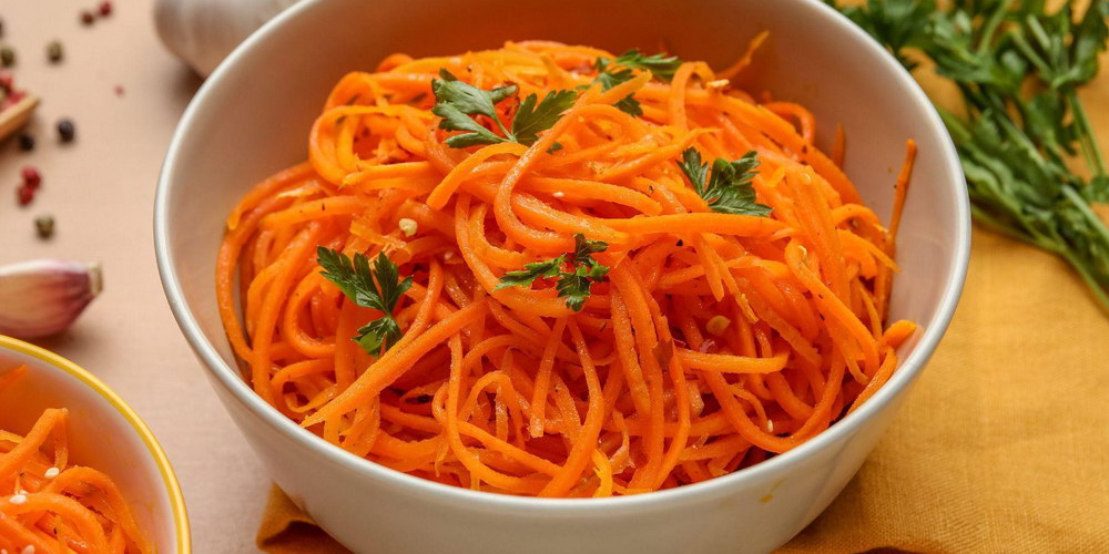 Морква по-корейськи — це салат, який давно став частиною домашньої кухні, хоча має зовсім не українське походження. Його готують на свята, додають до м’ясних страв і подають як самостійну закуску. Якщо ви шукаєте, як приготувати моркву по-корейськи так, щоб вона була хрумкою, ароматною та збалансованою за смаком, цей матеріал стане покроковим орієнтиром. Ми розберемо покроковий рецепт, пояснимо роль спецій і покажемо, як змінювати гостроту без втрати характеру страви. Також ви дізнаєтесь, як зберігати салат і чи підходить морква по-корейськи на зиму. У тексті є практичні поради, приклади та варіанти подачі. Структура побудована так, щоб ви могли не просто повторити рецепт моркви, а зрозуміти логіку маринаду. Це дозволить готувати стабільно смачно навіть без вагів і точних мір. Що таке морква по-корейськи Морква по-корейськи — це маринована морква з додаванням оцту, спецій та олії, яка має яскравий аромат і легку гостроту. Хоча назва натякає на корейське походження, страва виникла як адаптація корейських салатів до доступних інгредієнтів. Морква корейська готується просто, але вимагає розуміння балансу кислоти, солі та спецій. У домашніх умовах важливо правильно нарізати овоч і дати йому час промаринуватися. Корейська морква в домашніх умовах часто відрізняється від магазинної більш натуральним смаком і свіжістю. Салат підходить як гарнір до м’яса, картоплі або рису. Його готують заздалегідь, адже смак розкривається не одразу. Саме тому запит “морква по-корейськи” стабільно популярний серед тих, хто шукає простий і яскравий салат. Історія та походження Історія страви пов’язана з корейською діаспорою, яка адаптувала традиційні рецепти до доступних продуктів. Замість рідкісних овочів використовували моркву, що була доступною та дешевою. Так з’явилася морква по-корейськи у сучасному вигляді. Рецепт моркви швидко поширився завдяки простоті приготування. Страва не потребує термічної обробки, лише правильного маринування. З часом з’явилися варіації з різними спеціями для моркви. У кожному регіоні додають щось своє — більше часнику, гострого перцю або цукру. Сьогодні морква по-корейськи сприймається як класична закуска до багатьох страв. Чому вона така популярна Популярність пояснюється простотою та яскравим смаком. Якщо ви думаєте, як приготувати моркву смачно без складних інгредієнтів, цей варіант ідеальний. Морква корейська готується з доступних продуктів і не вимагає особливих навичок. Салат довго зберігається в холодильнику, що зручно для зайнятих людей. Його можна зробити гострим або більш м’яким за смаком. Крім того, смакова маринована морква по-корейськи добре поєднується з м’ясними стравами. Вона додає контрасту й текстури до звичних гарнірів. Саме універсальність робить її постійним гостем на столі. Інгредієнти для моркви по-корейськи Щоб морква по-корейськи вийшла збалансованою, важливо правильно підібрати інгредієнти моркви та пропорції. Основу складає свіжа соковита морква, оцет, рослинна олія та спеції для моркви. Деякі рецепти передбачають моркву по-корейськи з оцтом столовим або яблучним, що впливає на смак. Важливо не переборщити з кислотою, щоб салат не став занадто різким. Маринад для моркви по-корейськи повинен бути ароматним, але не домінувати над смаком овоча. Якщо морква солодка, кількість цукру можна зменшити. Рецепт моркви гнучкий, але баланс інгредієнтів має значення. Далі — базовий набір продуктів. Для класичного варіанту підготуйте такі інгредієнти: Свіжа морква — 1 кг Часник — 3–4 зубчики Оцет — 1–2 ст. ложки Рослинна олія — 3–4 ст. ложки Цукор — 1 ст. ложка Сіль — за смаком Червоний перець — за смаком Коріандр мелений Основні компоненти та пропорції Спеції для моркви формують характер страви, тому їх варто додавати поступово. Маринад для моркви по-корейськи повинен містити кислу складову, солодкість і ароматну частину. Рецепт моркви допускає варіації, але базові пропорції краще зберігати. Якщо ви додаєте більше оцту, компенсуйте це невеликою кількістю цукру. Морква по-корейськи з оцтом має виразніший смак, але не повинна бути різкою. Олію часто прогрівають і заливають на спеції, щоб аромат розкрився. Саме цей прийом дозволяє досягти глибшого смаку. Дотримання пропорцій — запорука стабільного результату. Покроковий рецепт моркви по-корейськи Покроковий рецепт допоможе уникнути помилок і отримати хрумку текстуру. Перед тим як приготувати салат, моркву потрібно натерти на спеціальній тертці. Це забезпечить правильну форму й маринування. Далі додаються інгредієнти моркви та спеції. Як приготувати моркву по-корейськи без зайвої гіркоти? Важливо не перетримати оцет і правильно змішати маринад. Смакова маринована морква по-корейськи розкривається через кілька годин. Краще дати їй настоятися мінімум 2–4 години. Так текстура стане м’якою, але залишиться хрумкою. Ось зрозуміла послідовність дій, щоб морква вийшла хрумкою: Натерти моркву на спеціальній тертці. Додати сіль і злегка перемішати. Додати цукор, оцет і спеції. Розігріти олію та залити на спеції. Додати подрібнений часник. Ретельно перемішати. Залишити маринуватися 2–6 годин. Морква по-корейськи найсмачніша після маринування: спеції “розкриваються”, а текстура стає хрумкою й соковитою. Секрети правильної моркви по-корейськи Морква по-корейськи вимагає точності в деталях. Спеції для моркви повинні бути свіжими, інакше аромат буде слабким. Маринад для моркви по-корейськи краще додавати поступово. Якщо ви сумніваєтесь, як приготувати збалансований варіант, почніть із мінімальної кількості оцту. Корейська морква в домашніх умовах виграє від часу настоювання. Не варто подавати її одразу після змішування. Також важливо використовувати якісну олію без стороннього запаху. Дрібні нюанси визначають фінальний смак. Як зробити ароматно та прибрати “запах сирої моркви” Щоб морква корейська не мала запаху сирого овоча, важливо правильно прогріти олію. Гарячу олію ллють на спеції для моркви, а не безпосередньо на овоч. Такий прийом дозволяє розкрити аромат коріандру та перцю. Якщо ви думаєте, як приготувати більш насичений варіант, додайте трохи паприки. Морква по-корейськи з оцтом має бути збалансованою, без різкого аромату. Додатковий часник посилює смак. Усе залежить від ваших уподобань. Тонкощі нарізки та маринування Покроковий рецепт завжди починається з правильної нарізки. Морква повинна бути довгою й тонкою. Корейська морква в домашніх умовах потребує рівномірного перемішування. Час маринування впливає на текстуру. Якщо залишити салат на ніч, смак стане глибшим. Гостра морква по-корейськи з часником набуває насиченого аромату через кілька годин. Не поспішайте з подачею. Дайте спеціям “попрацювати”. Маринад — це серце цього салату: правильний баланс кислоти, солі та спецій задає весь смак. Варіації приготування Рецепт моркви можна змінювати залежно від уподобань. Морква по-корейськи добре поєднується з іншими овочами. Деякі додають болгарський перець або кунжут. Гостра морква по-корейськи з часником підходить для любителів пікантного смаку. Якщо зменшити оцет, салат стане м’якшим. Морква по-корейськи на зиму готується з трохи більшою кількістю кислоти. Варіації дозволяють створити власний рецепт. Головне — не втрачати баланс. Морква по-корейськи з часником і гостринкою Гостра морква по-корейськи з часником має насичений аромат. Для цього додають більше червоного перцю та свіжого часнику. Морква по-корейськи з оцтом у цьому варіанті повинна бути збалансованою. Як приготувати без надмірної гостроти? Додайте трохи цукру для пом’якшення. Спеції для моркви краще обсмажити сухими перед додаванням. Це посилить аромат. Варіант ідеально підходить до м’яса. Як зберігати моркву по-корейськи Питання, як зберігати моркву по-корейськи, важливе для тих, хто готує великими порціями. Салат тримають у холодильнику в щільно закритій тарі. Морква по-корейськи на зиму готується з більшою кількістю оцту. Якщо ви плануєте зберігати довше трьох днів, використовуйте скляну банку. Як зберігати правильно? Не залишайте салат при кімнатній температурі надовго. Волога й тепло скорочують термін придатності. Спосіб Тара Температура Термін Холодильник Скляна банка +4°C 3–5 днів На зиму Стерилізована банка +4°C До 2 тижнів Поради й часті помилки Морква по-корейськи може не вийти, якщо порушити баланс спецій. Спеції для моркви потрібно додавати поступово. Морква по-корейськи з оцтом не повинна бути надто кислою. Якщо ви пересолили, додайте трохи цукру. Якщо салат занадто м’який, зменште час маринування. Рецепт моркви простий, але потребує уваги до деталей. Дотримуйтесь пропорцій. І не поспішайте з подачею. Ось помилки, через які салат виходить “плаский” або занадто кислий: Занадто багато оцту. Недостатньо спецій. Неякісна олія. Мало часу для маринування. Неправильна нарізка. Як подавати — ідеї для подачі та комбінації страв Морква по-корейськи універсальна в подачі. Вона підходить до м’яса, риби та овочевих страв. Якщо ви шукаєте, як приготувати моркву смачно для святкового столу, додайте зелень і кунжут. Салат добре виглядає в прозорому посуді. Його можна використовувати як інгредієнт для рулетів або сендвічів. Морква по-корейськи додає контрасту до гарнірів. Вона підходить навіть до простого рису. Спробуйте експериментувати. Спробуйте такі поєднання, якщо хочете урізноманітнити подачу: З курячим філе У лаваші З печеною картоплею Як начинка для рулетів З рисом або булгуром Цей салат працює як універсальна добавка: він додає гостроти, свіжості й “характеру” навіть простим стравам.